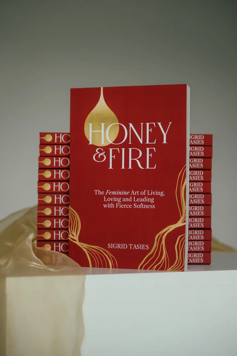 A copy of Honey & Fire standing upright on a white plinth, a stack of copies behind it and gold organza draped at its base